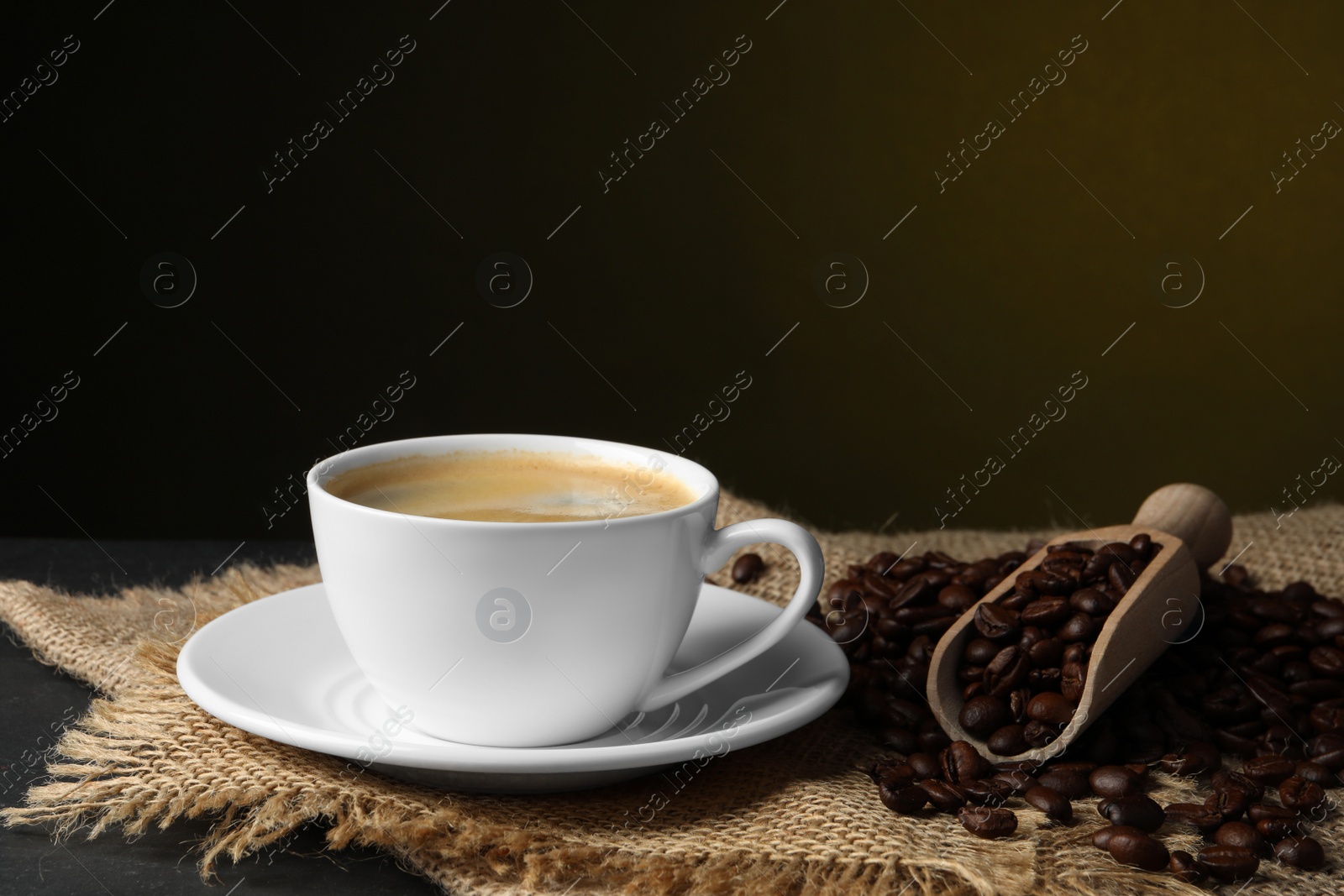 Photo of Cup of hot aromatic coffee and scoop with roasted beans on black table against dark background