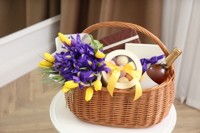 Wicker basket with gift, bouquet and wine on white table indoors