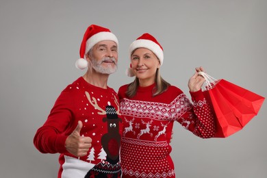 Senior couple in Christmas sweaters and Santa hats with shopping bags on grey background