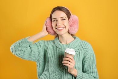 Happy woman with cup of drink wearing warm earmuffs on yellow background