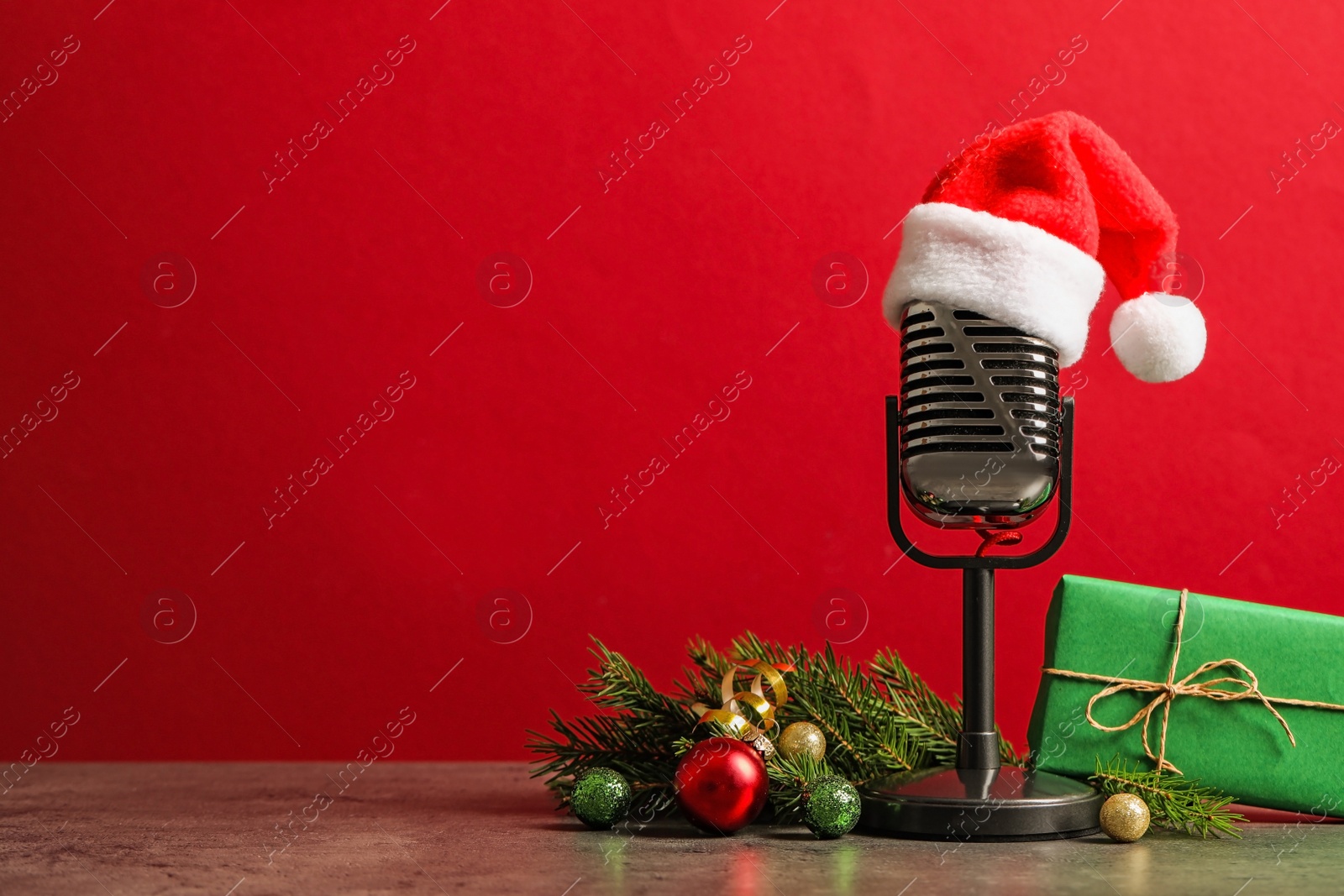 Photo of Microphone with Santa hat and decorations on grey table against red background, space for text. Christmas music