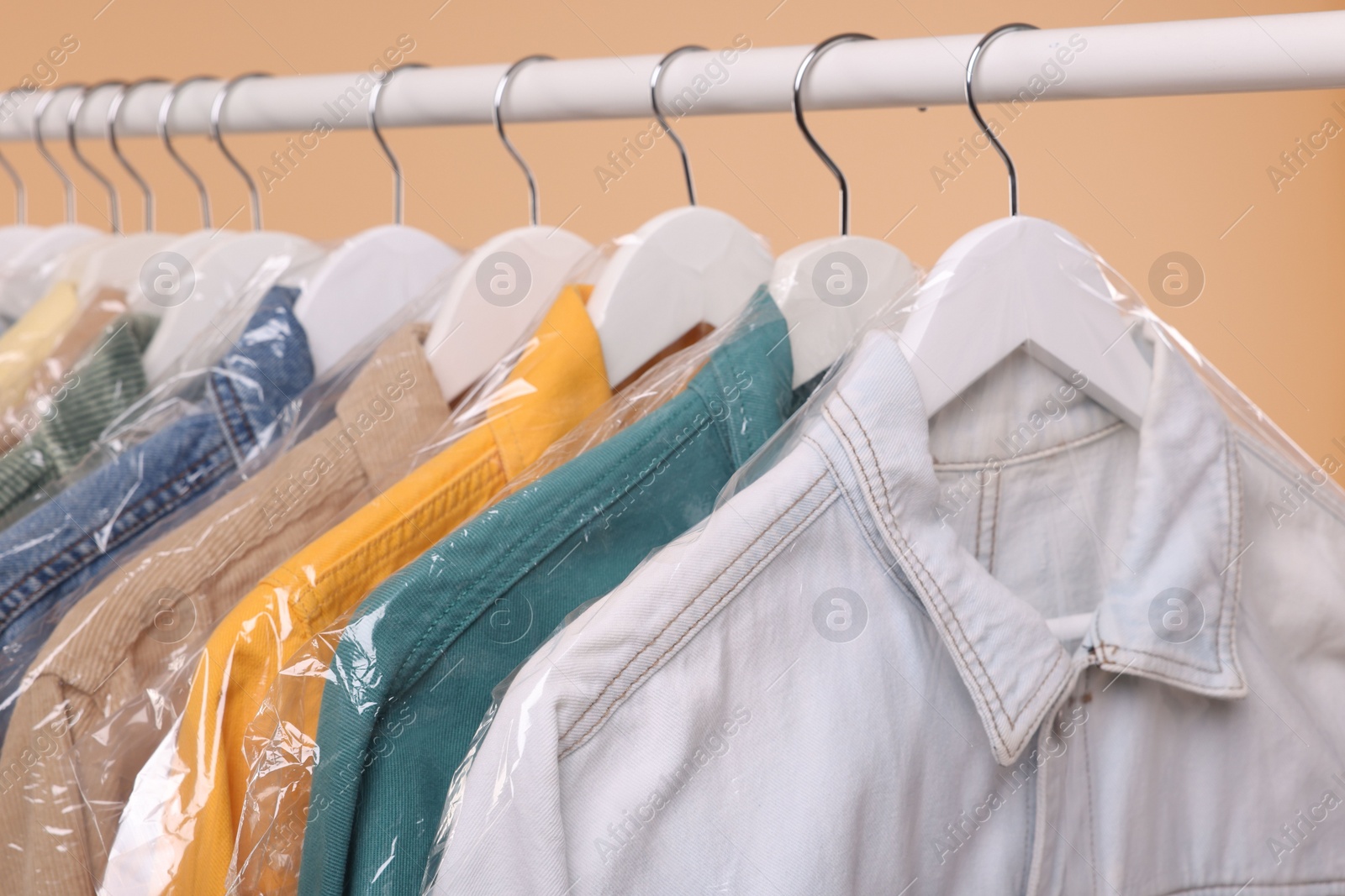 Photo of Dry-cleaning service. Many different clothes in plastic bags hanging on rack against beige background, closeup