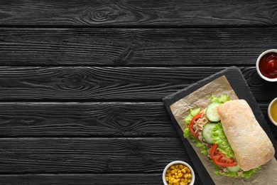 Photo of Food photography. Delicious sandwich with tuna, vegetables and sauces on black wooden table, flat lay with space for text