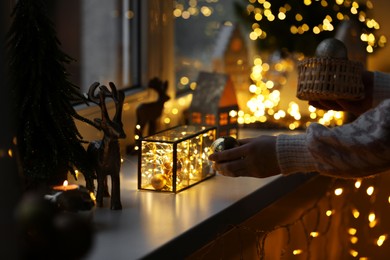 Photo of Christmas atmosphere. Woman holding beautiful baubles near window sill indoors, closeup