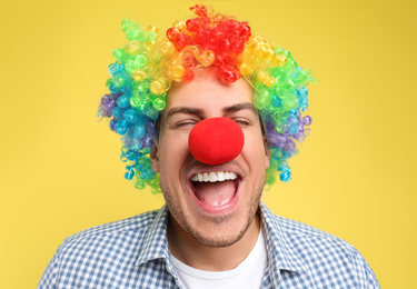 Photo of Funny man with clown nose and rainbow wig on yellow background. April fool's day