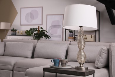 Photo of Comfortable sofa in living room, focus on table with cups and lamp