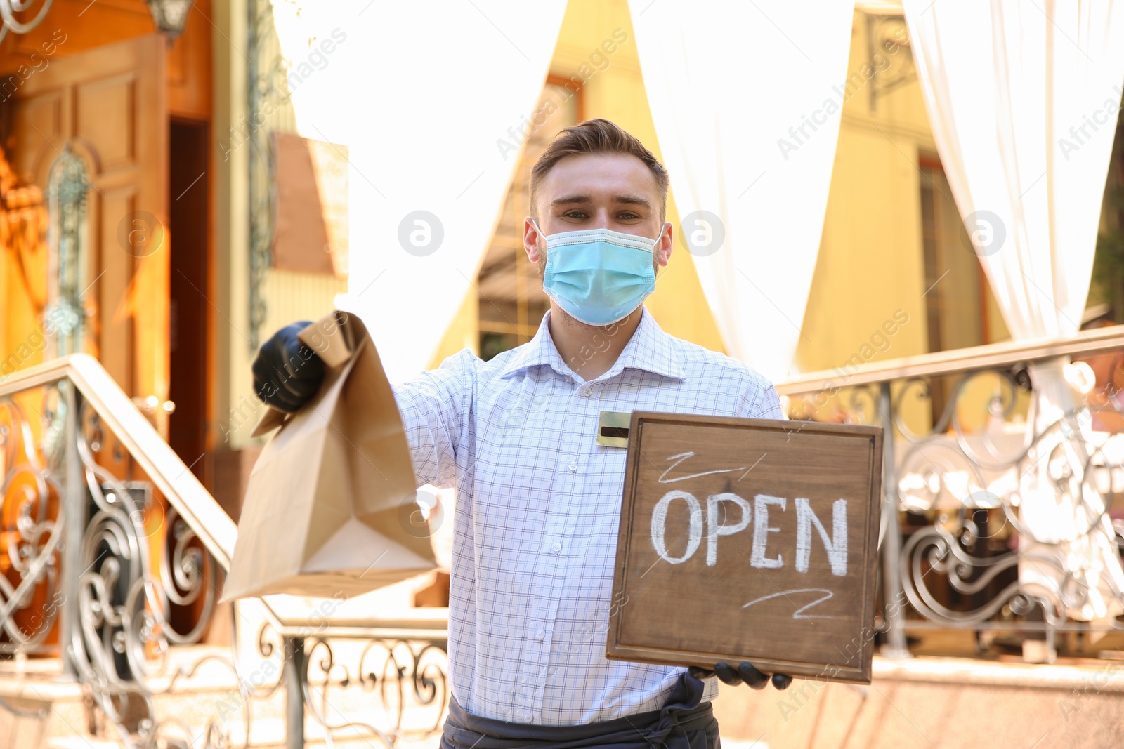 Photo of Waiter with packed takeout order and OPEN sign near restaurant. Food service during coronavirus quarantine