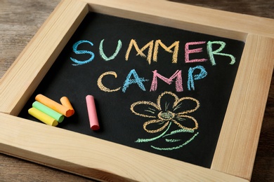 Photo of Small blackboard with text SUMMER CAMP, drawing and chalk sticks on wooden background, closeup