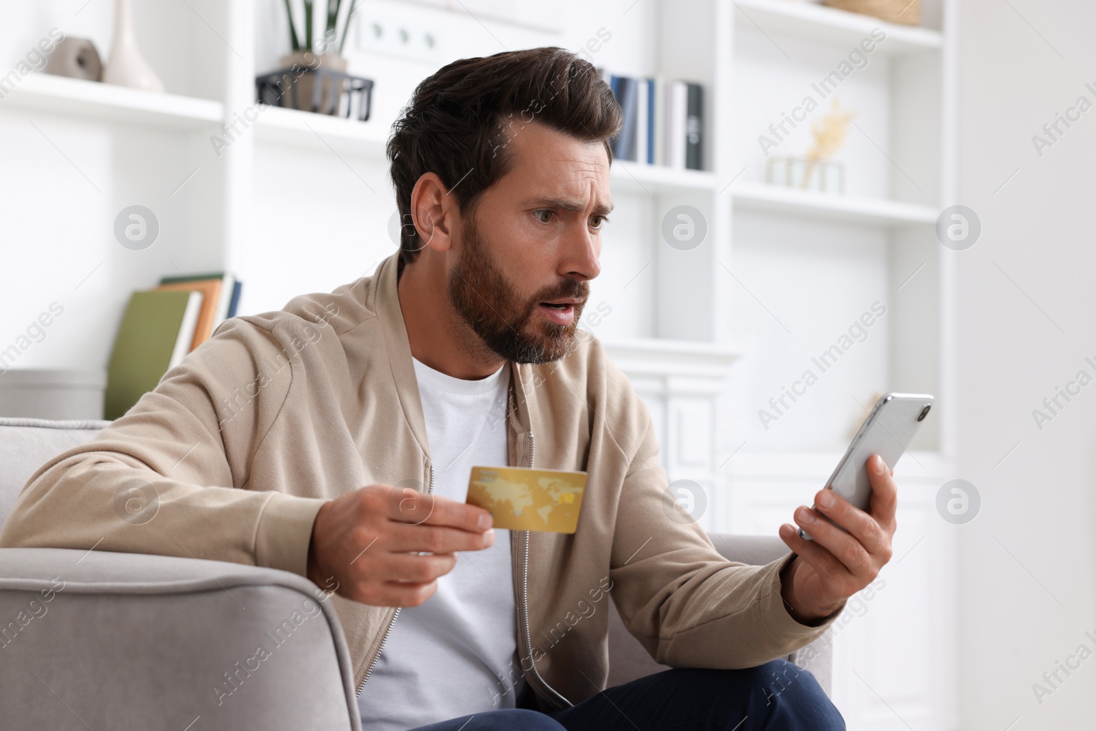 Photo of Upset man with credit card using smartphone in armchair at home. Be careful - fraud