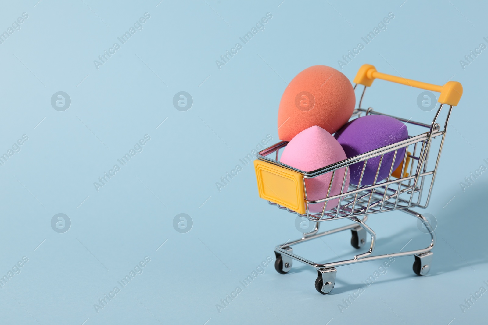 Photo of Makeup sponges in small shopping cart on light blue background. Space for text