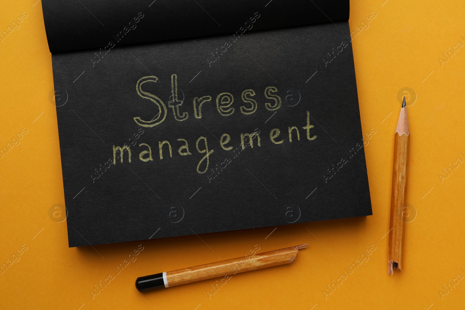 Photo of Notebook with text Stress Management and broken pencil on orange background, flat lay