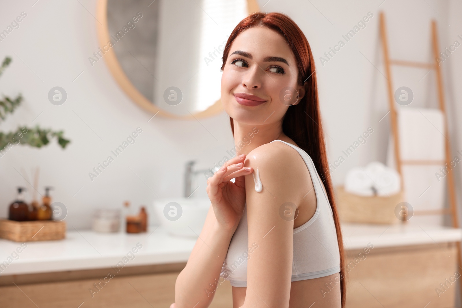 Photo of Beautiful young woman applying body cream onto shoulder in bathroom, space for text