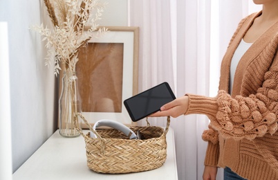 Photo of Woman putting smartphone into wicker basket with gadgets at home. Digital detox concept