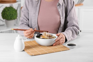 Woman eating delicious ramen with chopsticks at white table indoors, closeup. Noodle soup