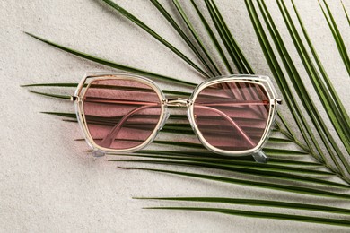Photo of Stylish sunglasses and palm leaf on sand, top view