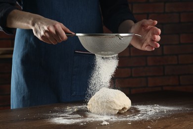 Photo of Making bread. Woman sprinkling flour onto dough at wooden table indoors, closeup