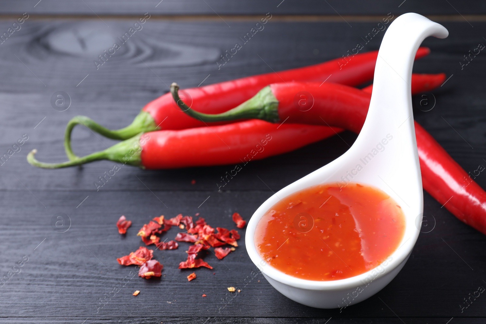 Photo of Spicy chili sauce in spoon and peppers on black wooden table, closeup