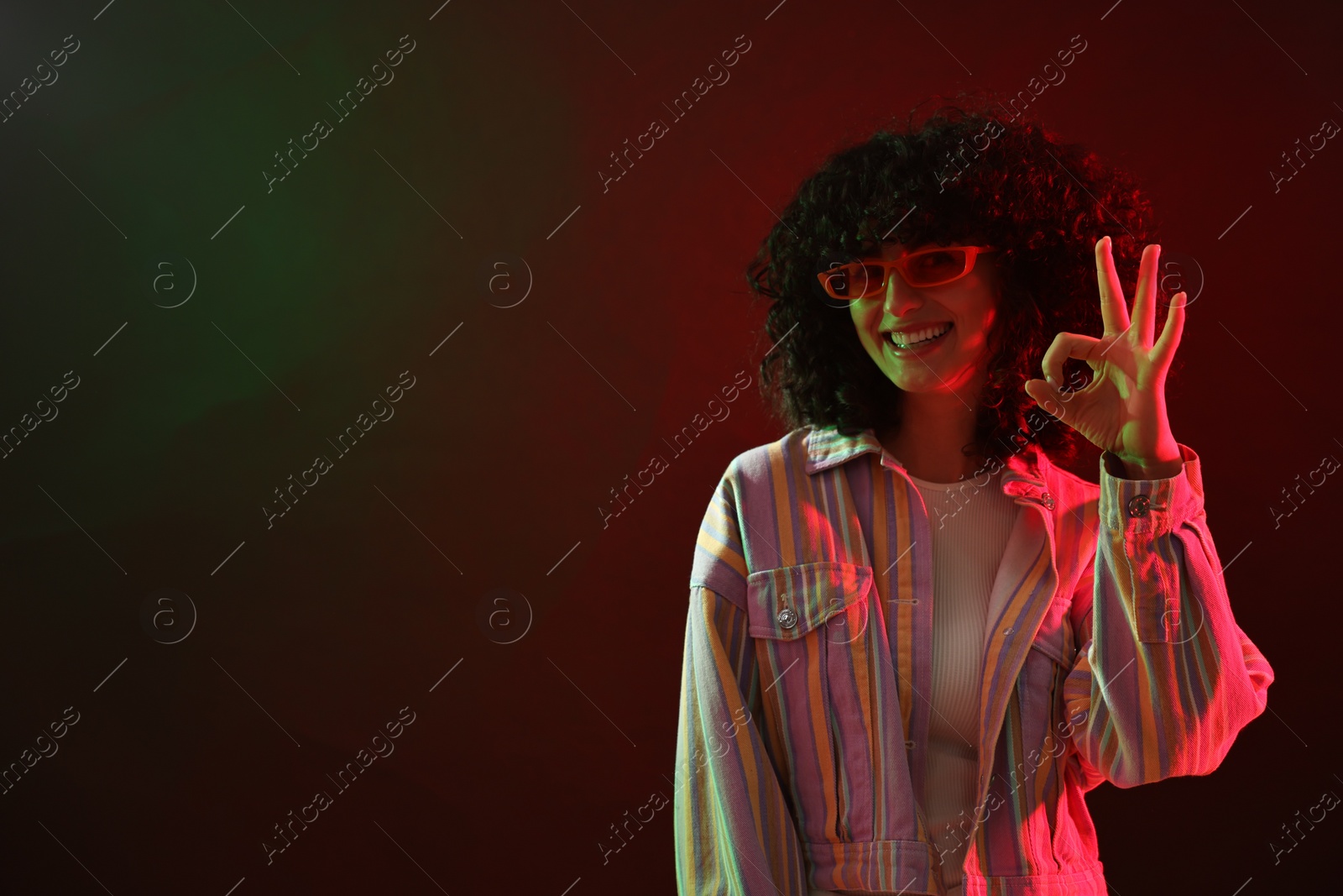 Photo of Beautiful young woman in sunglasses making ok gesture on color background in neon lights