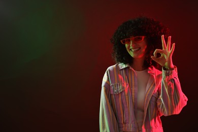 Photo of Beautiful young woman in sunglasses making ok gesture on color background in neon lights