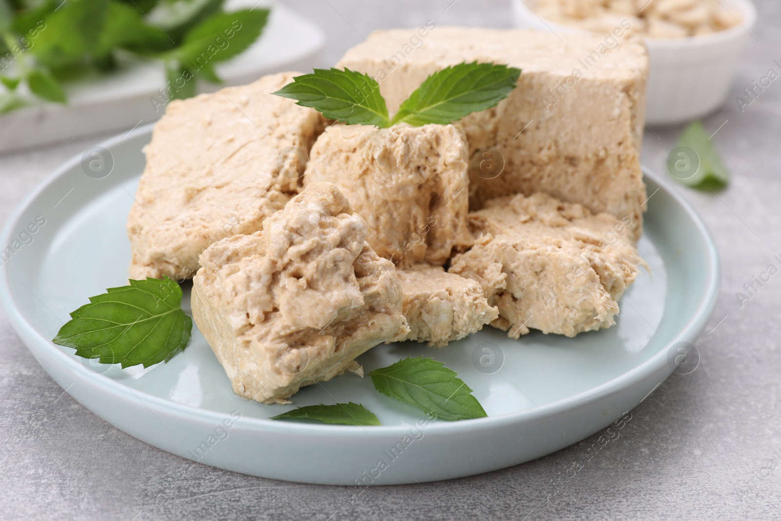 Photo of Plate with pieces of tasty halva and mint leaves on light grey table, closeup