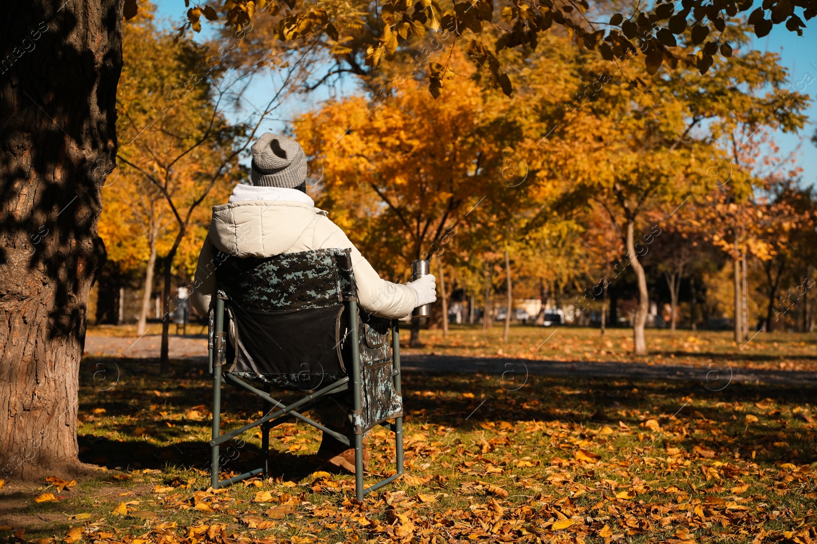 Photo of Woman with thermos sitting in camping chair outdoors on autumn sunny day, back view