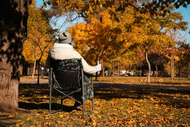 Woman with thermos sitting in camping chair outdoors on autumn sunny day, back view