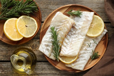 Photo of Fresh raw cod fillets with rosemary, lemon and oil on wooden table, flat lay