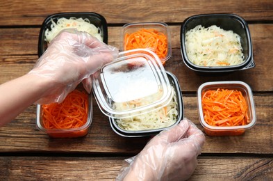 Waiter in gloves closing containers with salads at wooden table, closeup. Food delivery service