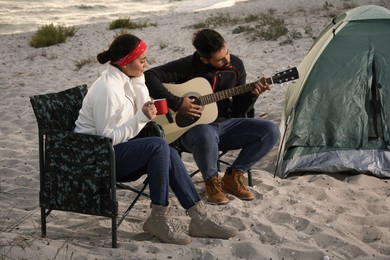 Photo of Young man playing guitar to his beloved girlfriend near camping tent on beach