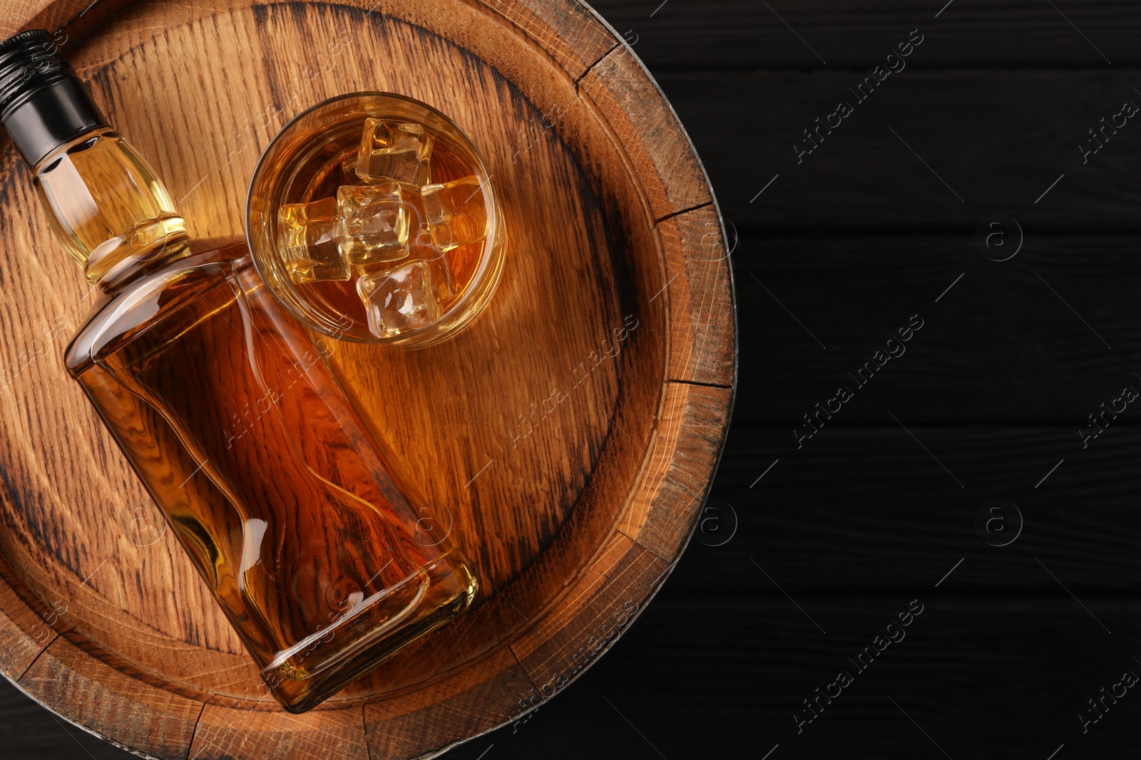 Photo of Whiskey with ice cubes in glass, bottle and barrel on black wooden table, top view. Space for text