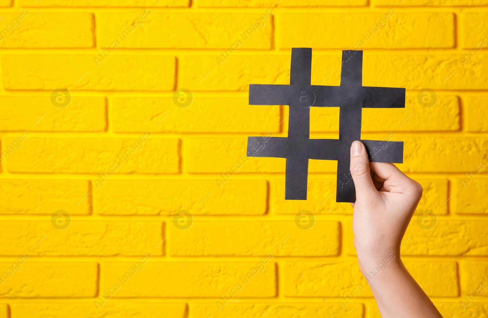 Photo of Woman holding black paper cutout symbol of hashtag on yellow brick wall, closeup. Space for text