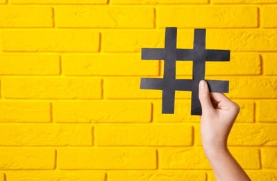 Woman holding black paper cutout symbol of hashtag on yellow brick wall, closeup. Space for text