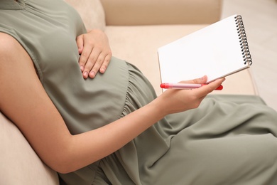 Pregnant woman with notebook and marker choosing name for baby on sofa, closeup