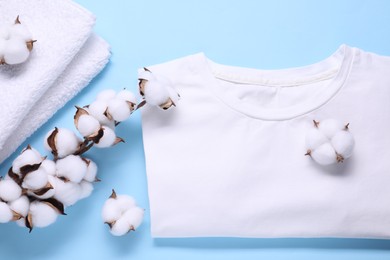 Photo of Cotton branch with fluffy flowers, white t-shirt and terry towel on light blue background, flat lay