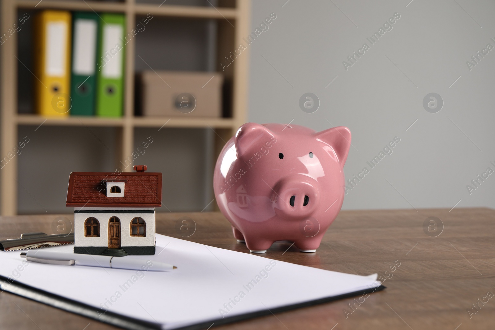 Photo of House model, piggy bank, clipboard and pen on wooden table indoors