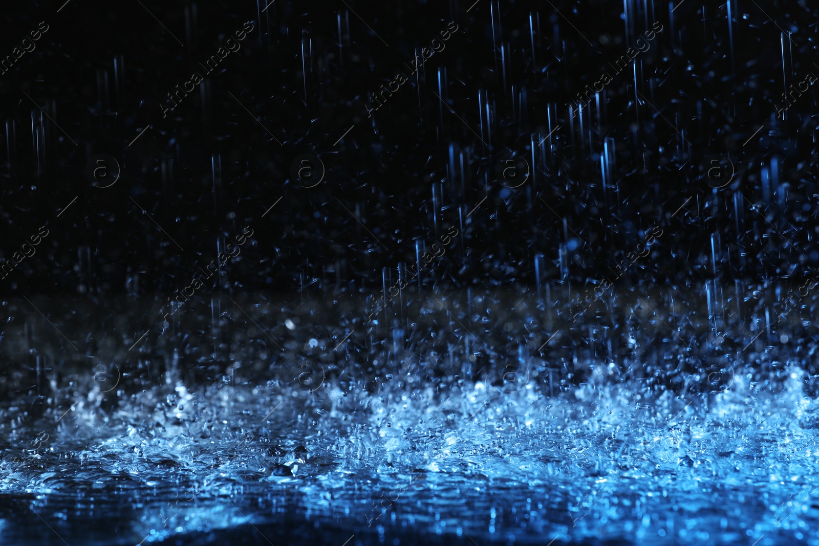 Photo of Heavy rain falling down on ground against dark background, toned in blue