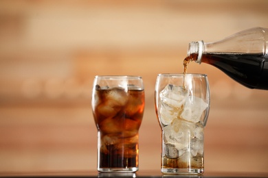 Pouring cola from bottle into glass with ice cubes on table against blurred background