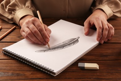 Woman drawing feather with graphite pencil in sketchbook at wooden table, closeup