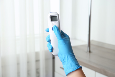 Photo of Doctor in gloves holding non contact infrared thermometer indoors, closeup