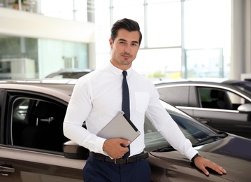 Young salesman with tablet near car in modern dealership