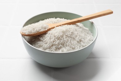 Photo of Raw basmati rice in bowl and spoon on white tiled table