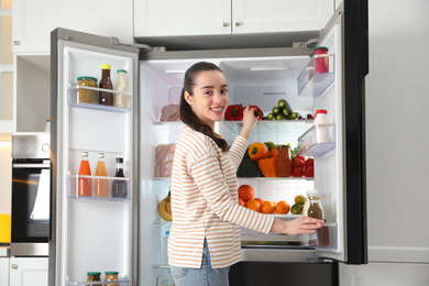 Photo of Young woman taking bell pepper out of refrigerator in kitchen