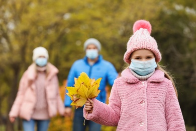 Photo of Cute girl in medical mask walking with her parents outdoors on autumn day. Protective measures during coronavirus quarantine
