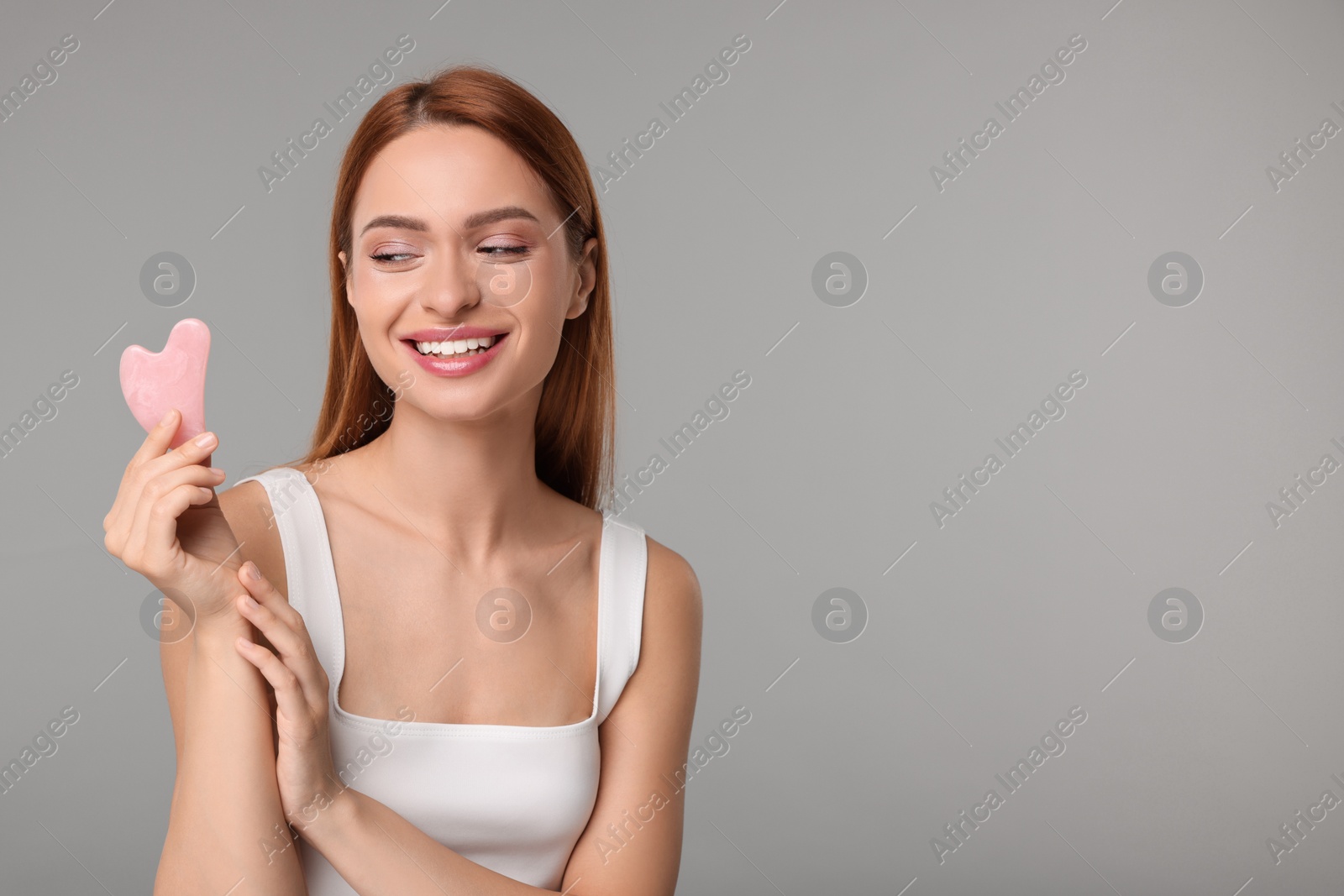 Photo of Young woman with rose quartz gua sha tool on grey background, space for text. Facial massage