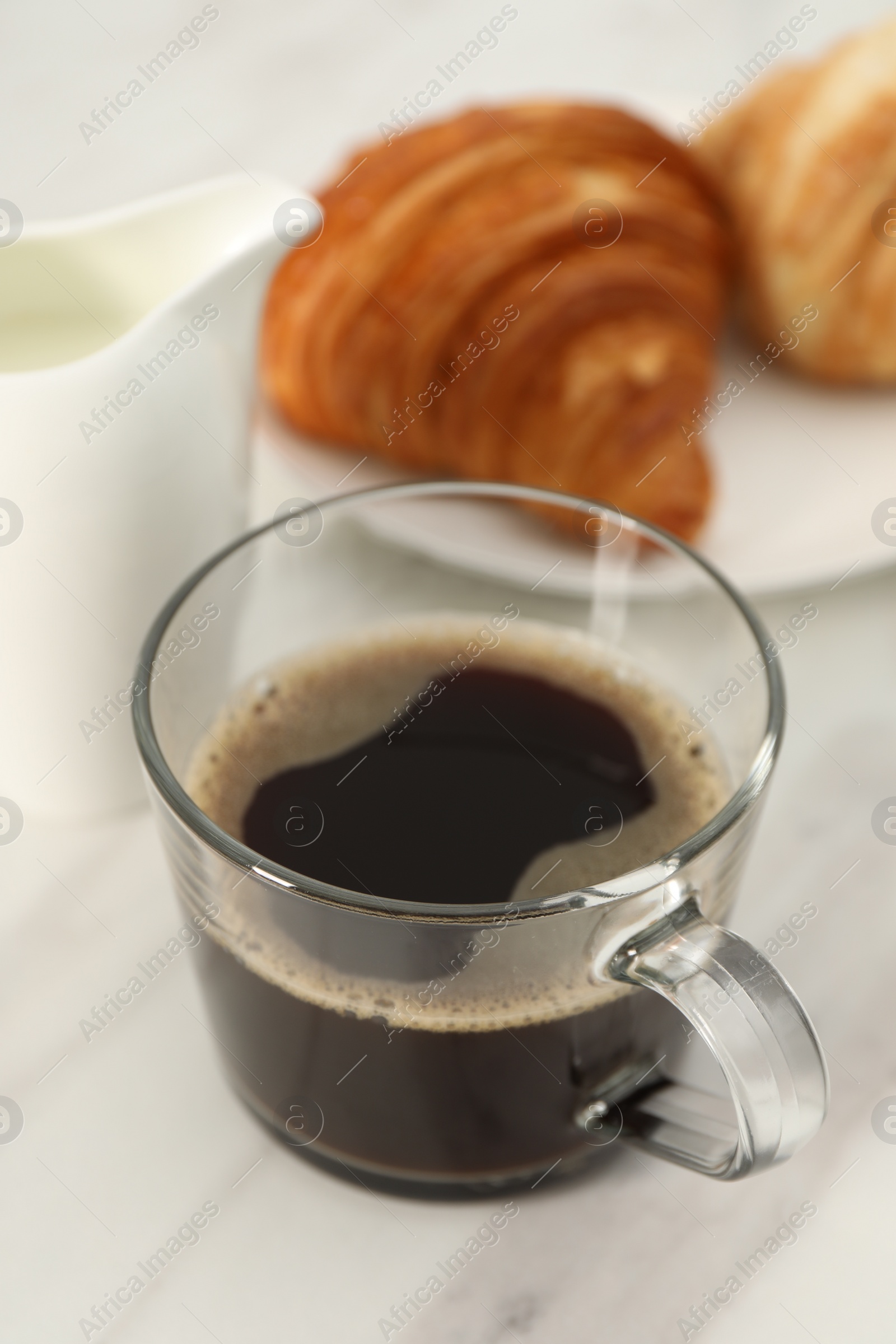 Photo of Aromatic coffee in glass cup, pitcher and fresh croissants on white marble table, closeup