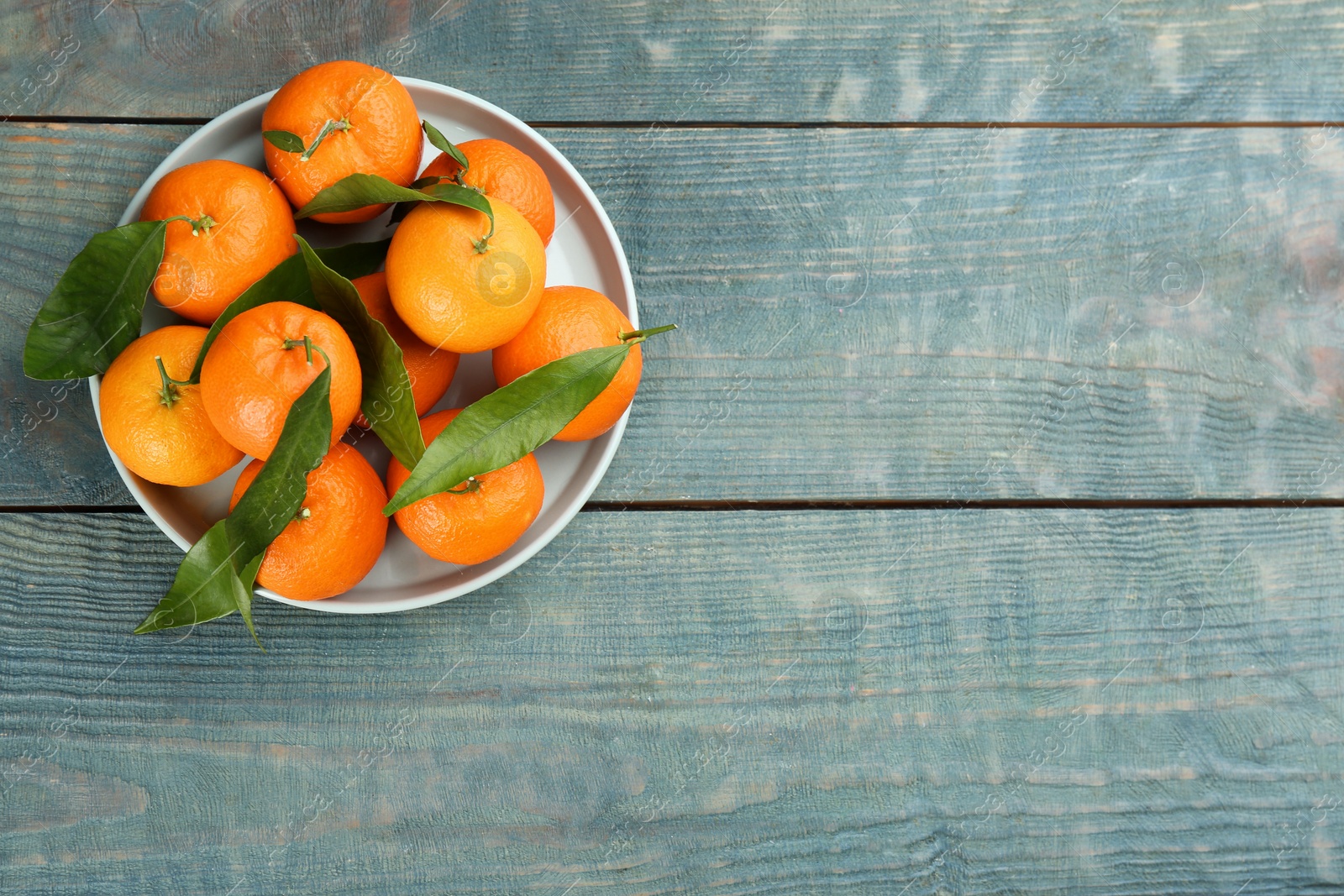 Photo of Fresh ripe tangerines with green leaves on blue wooden table, top view. Space for text