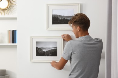 Photo of Young man hanging picture frames on white wall indoors, back view