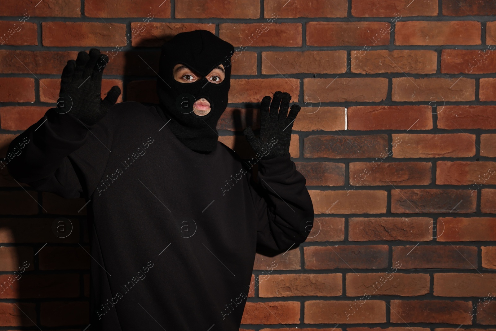 Photo of Thief in balaclava raising hands against red brick wall. Space for text