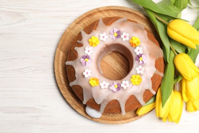 Photo of Delicious Easter cake decorated with sprinkles near beautiful tulips on white wooden table, flat lay. Space for text