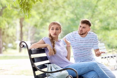 Photo of Young couple arguing while sitting on bench in park. Problems in relationship